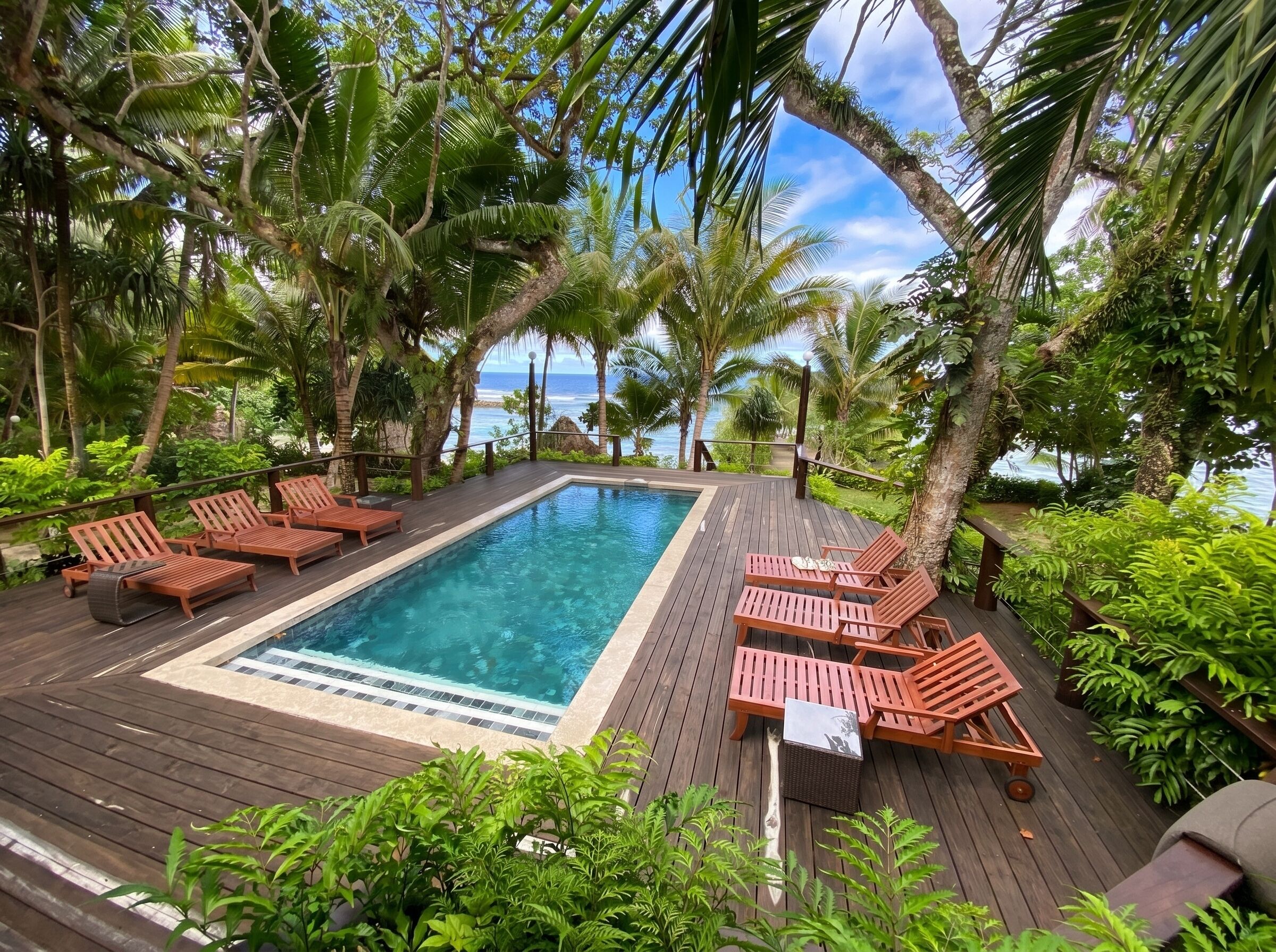 Una terraza de madera con piscina, sillas de descanso y vegetación tropical.