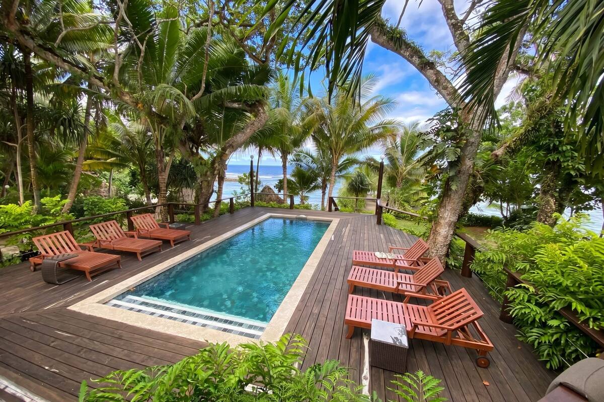 Eine Holzterrasse mit Pool, Liegestühlen und tropischer Bepflanzung.