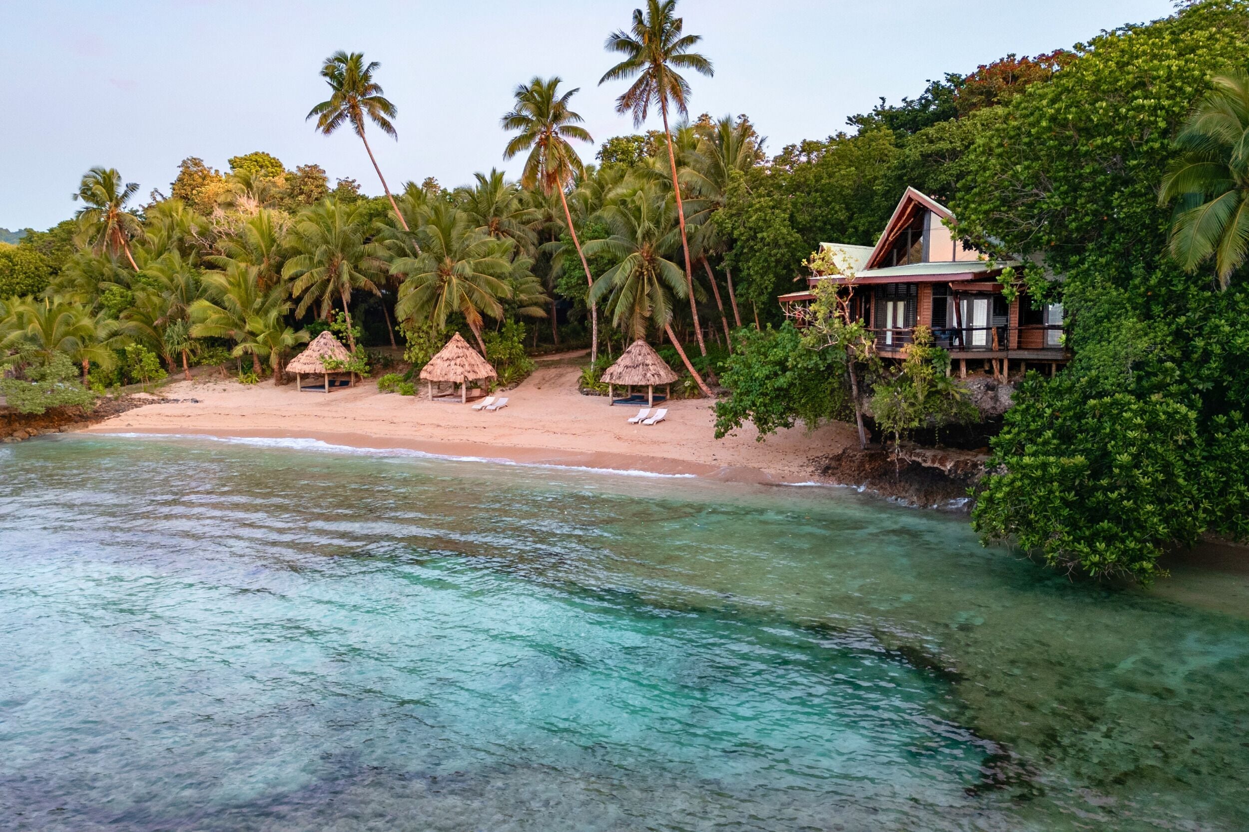 A beach with clear turquoise water, palm trees, and a wooden house with a balcony.