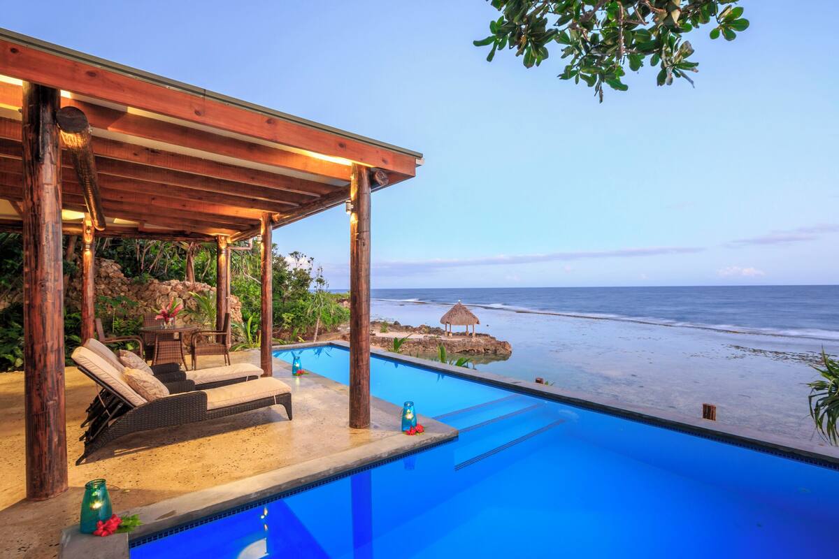 Ein Pool mit Liegestühlen und einer Holzpergola mit Blick auf einen Strand.