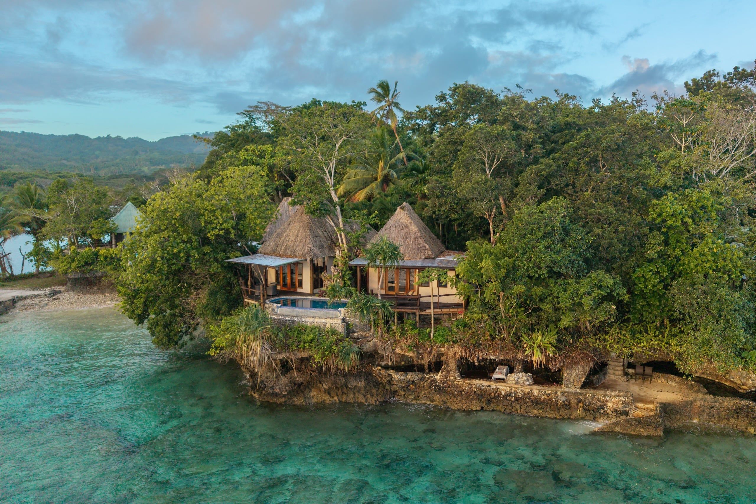 Un complexe hôtelier tropical situé sur une île, avec des bungalows aux toits de chaume, une piscine et une végétation luxuriante.