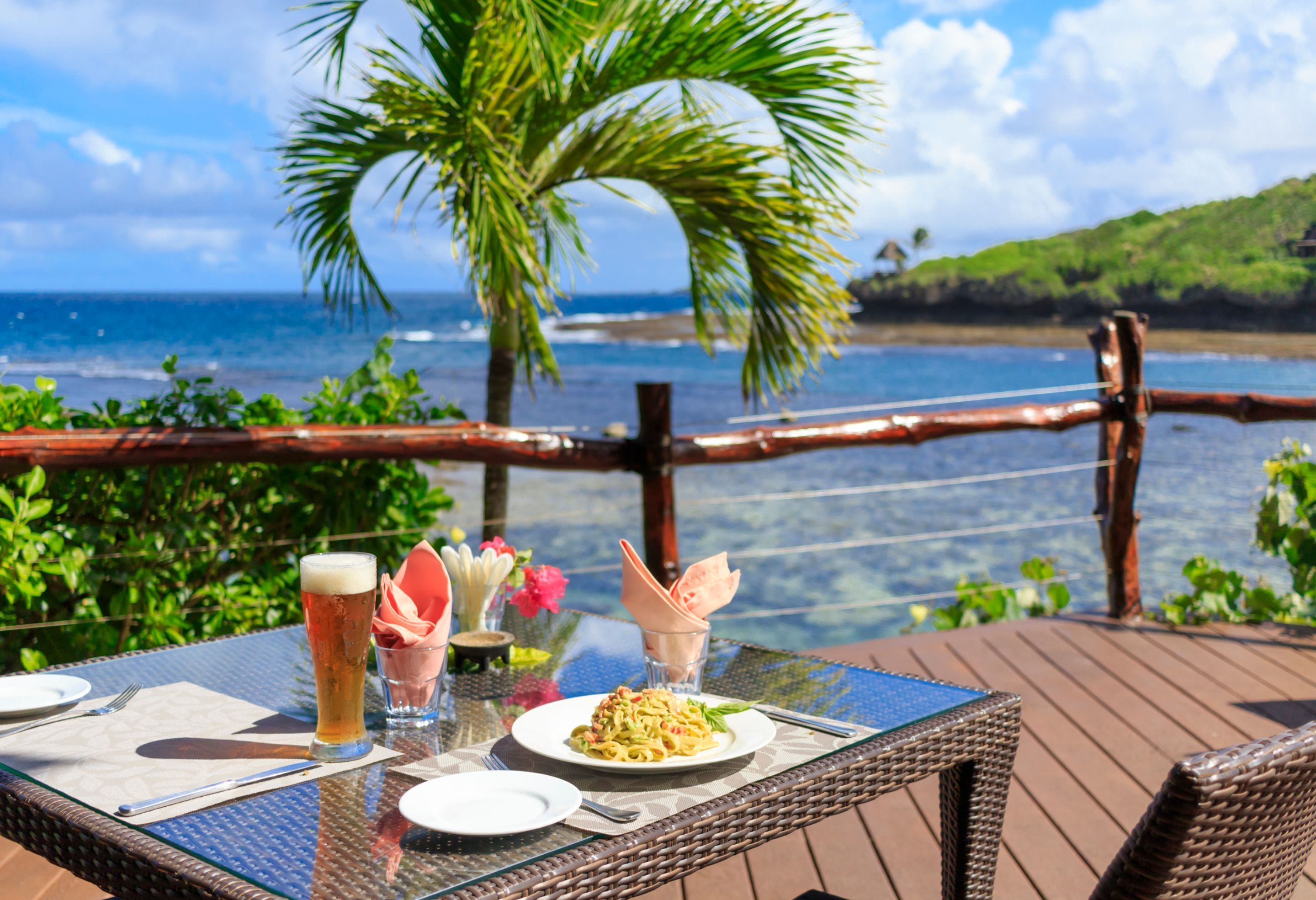 A dining table with a glass of beer, a plate of pasta, and a glass of water on a deck overlooking the ocean.