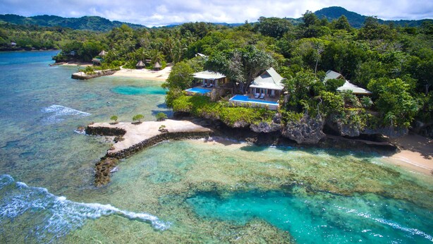 An aerial view of a tropical island with clear turquoise waters, a sandy beach, and several thatched-roof structures surrounded by lush greenery. - Savasi Island Resort (Savusavu)