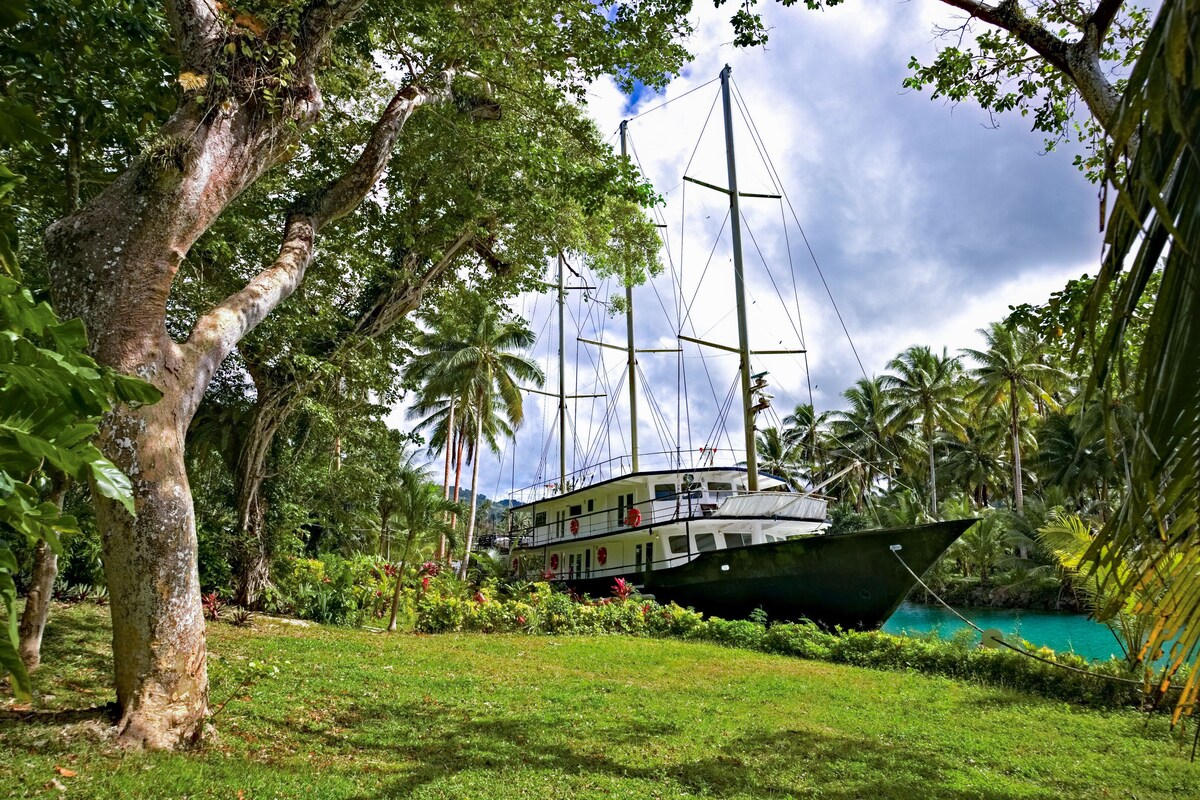 A boat docked in a lush, tropical garden with palm trees and a clear blue pool.