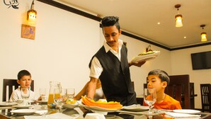 Un comedor con una mesa dispuesta para una comida, un camarero sirviendo y un niño sentado a la mesa.