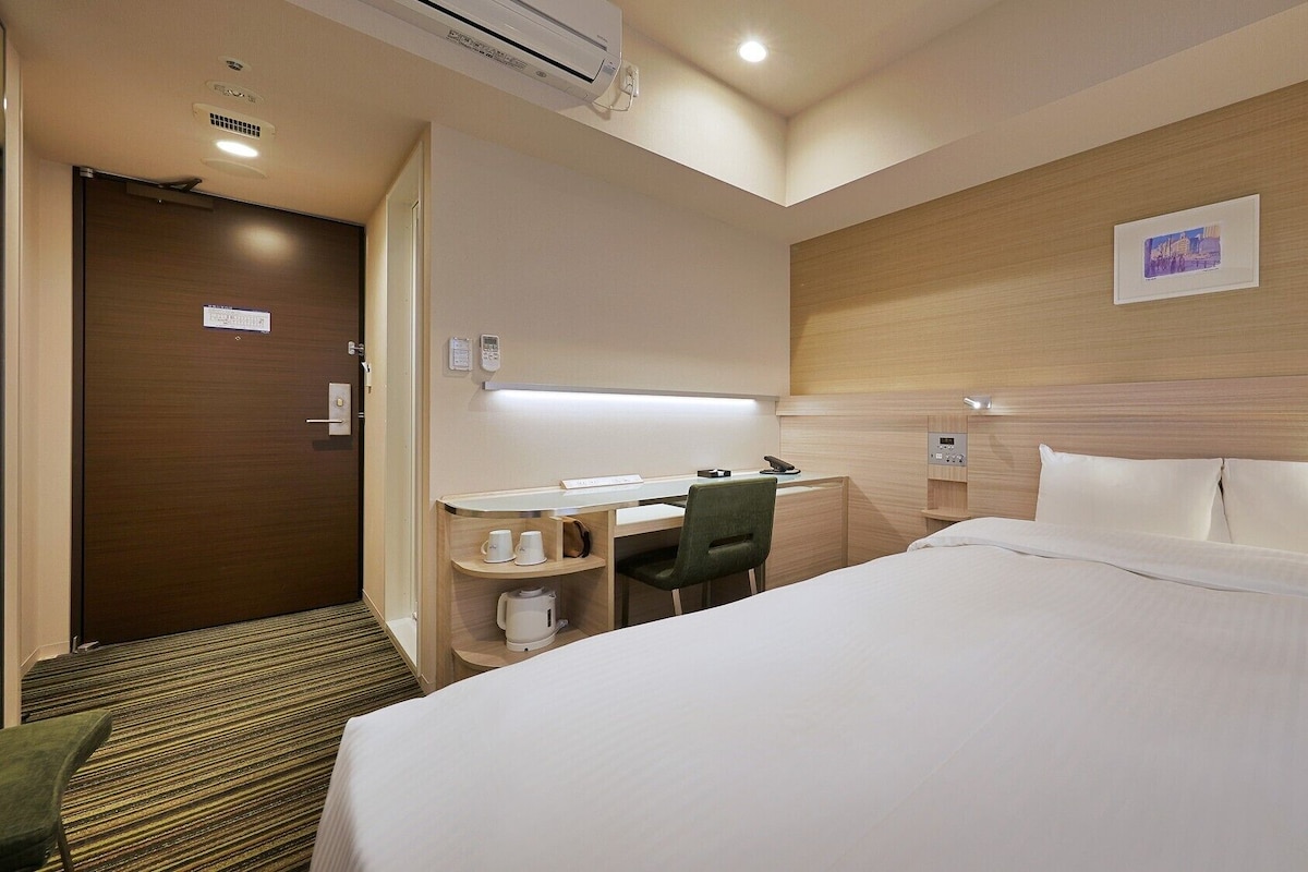 A hotel room with a bed, desk, chair, and a small kitchenette area.
