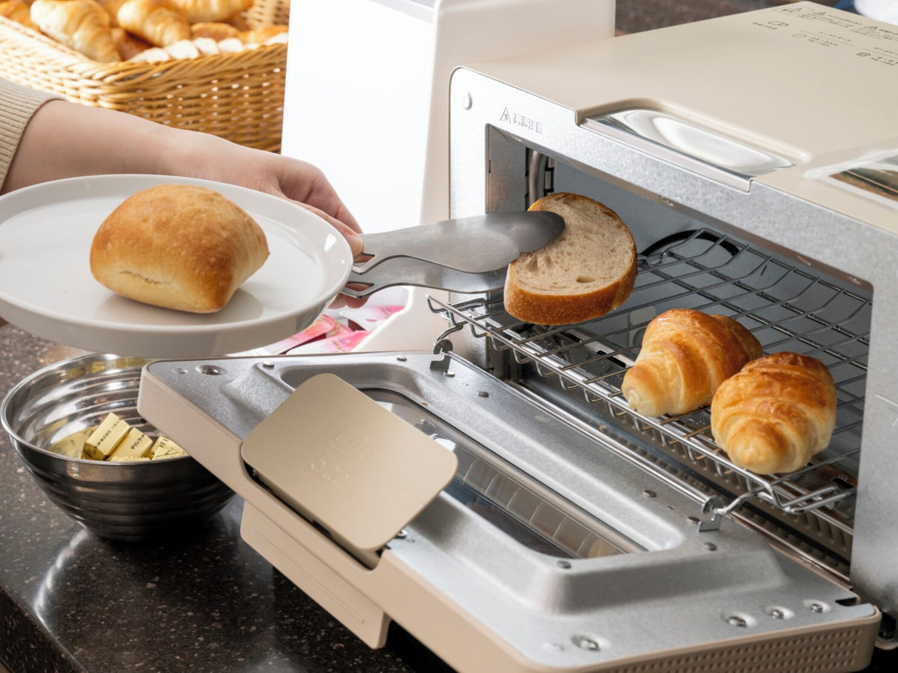 A toaster oven with bread and croissants inside, a plate with a roll, and a bowl of butter.