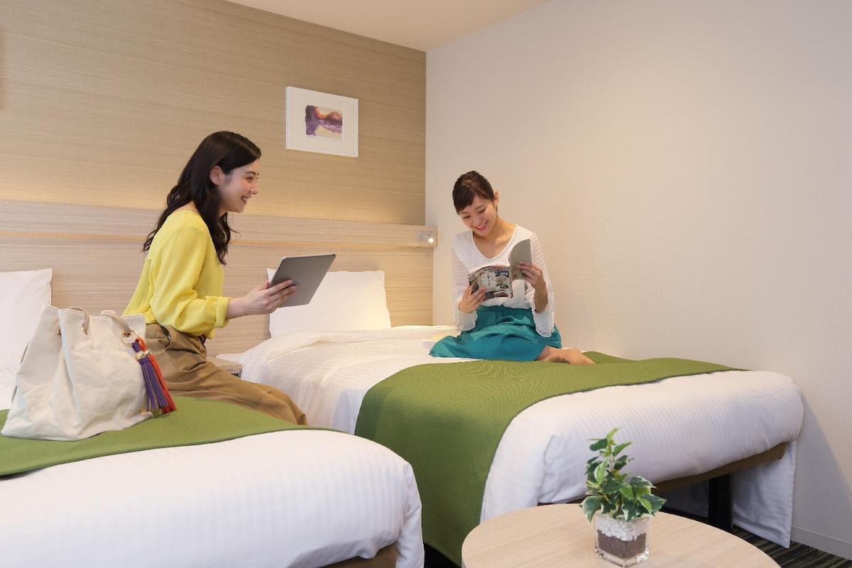 Two beds with green and white bedding, a small round table with a plant, and a wall-mounted picture.