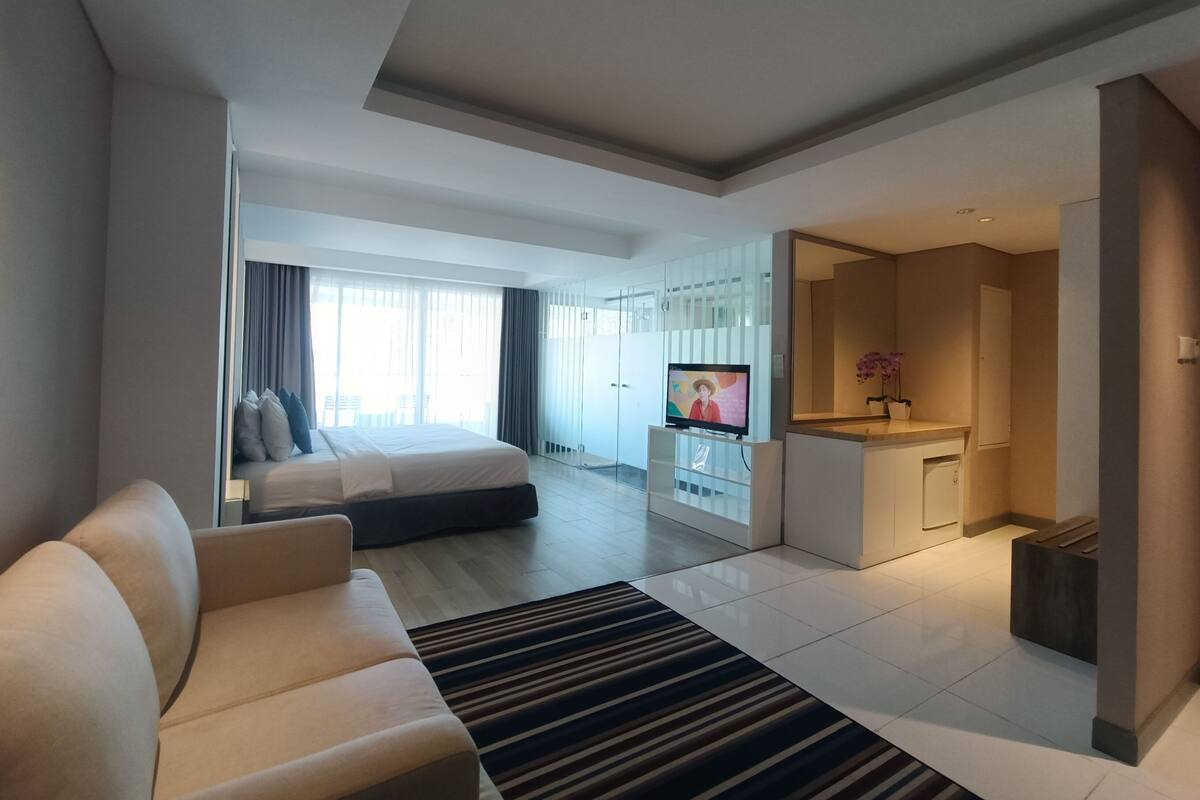 Ein modernes Hotelzimmer mit einem großen Bett, einer Sitzecke, einem Fernseher, einer Küchenzeile und einem Balkon mit Vorhängen.