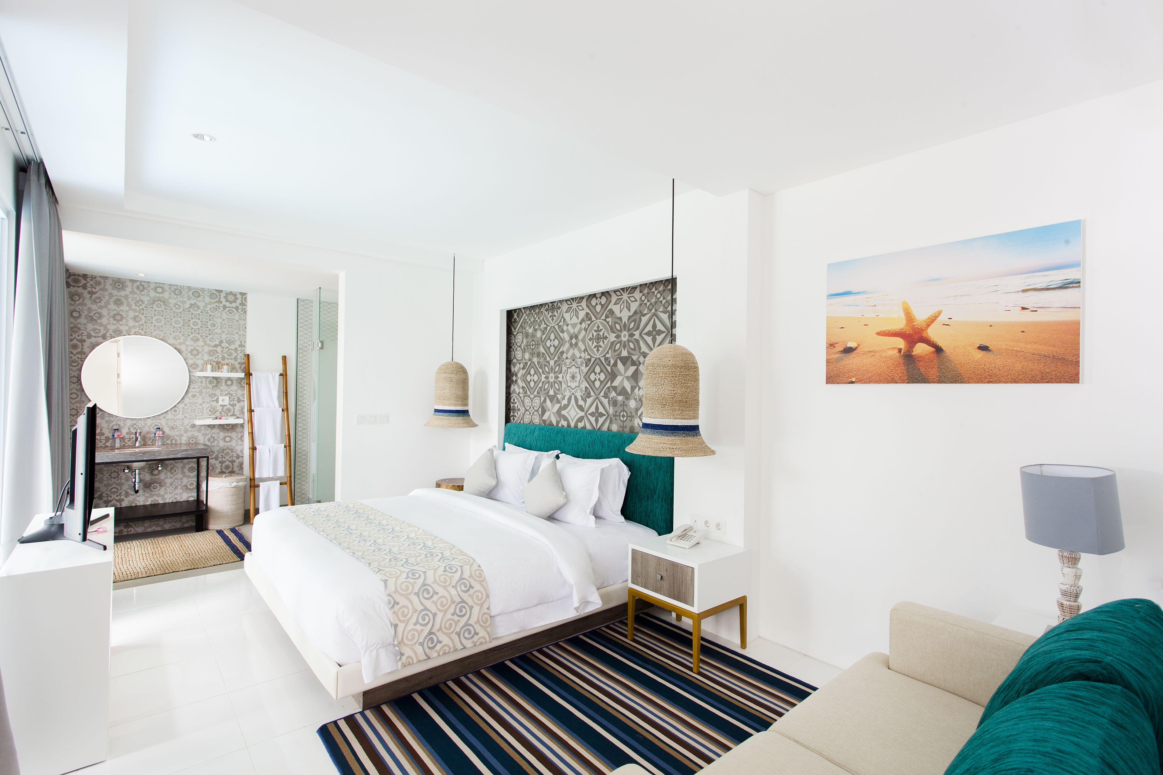 A modern hotel room with a large bed, a bedside table, a nightstand, a lamp, a painting of a beach, and a view of the bathroom.