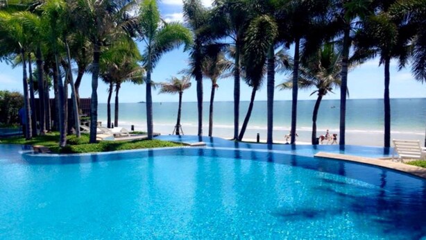 A clear blue pool surrounded by palm trees, with a beach and ocean in the background. - Baan San Ploen Condominium by Renvio (Hua Hin)