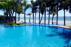 Ein klares, blaues Schwimmbecken, umgeben von Palmen, mit Strand und Meer im Hintergrund.