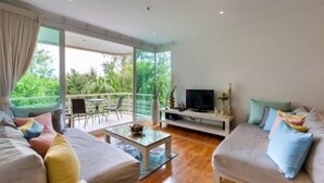 A spacious living room with a large window, a sofa set, a coffee table, and a TV on a stand. - Baan San Ploen Condominium by Renvio (Hua Hin)