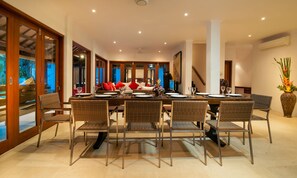 A dining area with a long table set for multiple people, surrounded by wicker chairs, with a view of a blue sky through large windows. - The Jasmina Estate (Seminyak)