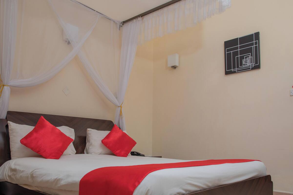 A hotel room with a bed, white curtains, red pillows, and a wall-mounted artwork.