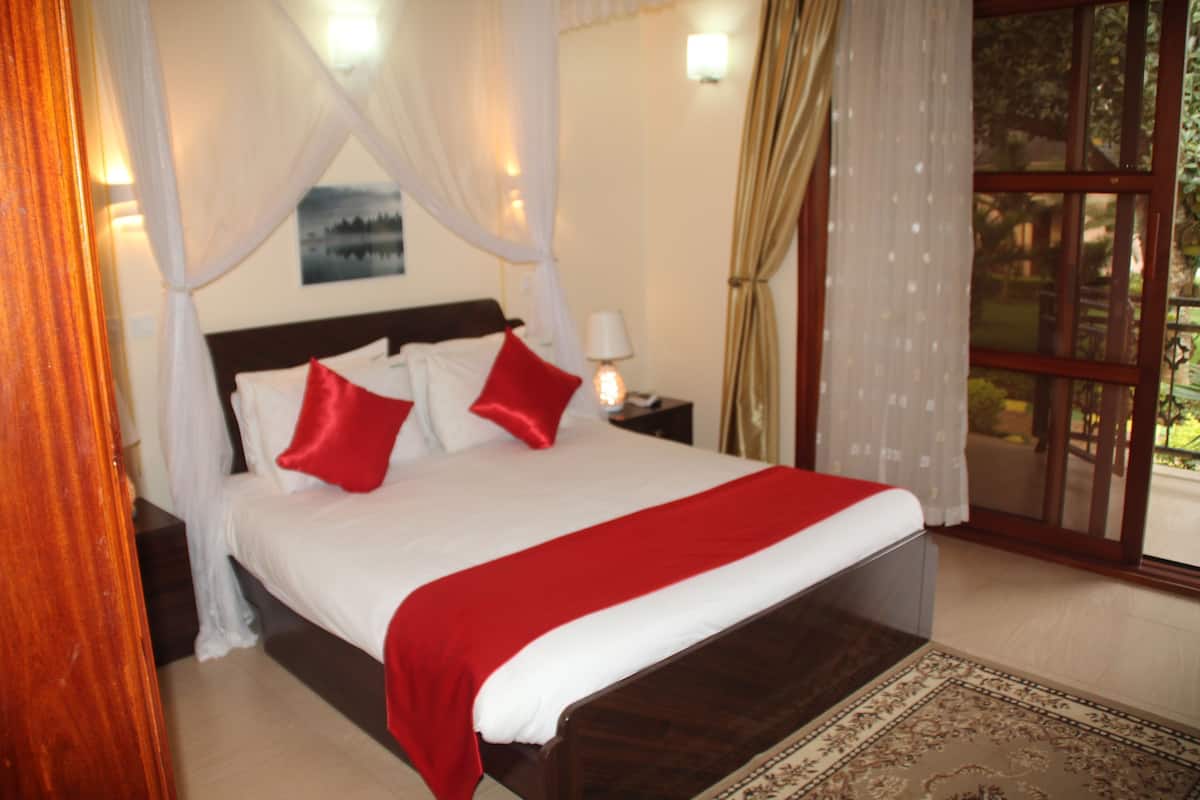 A bedroom with a bed, a nightstand, a lamp, a window with curtains, and a view of greenery outside.