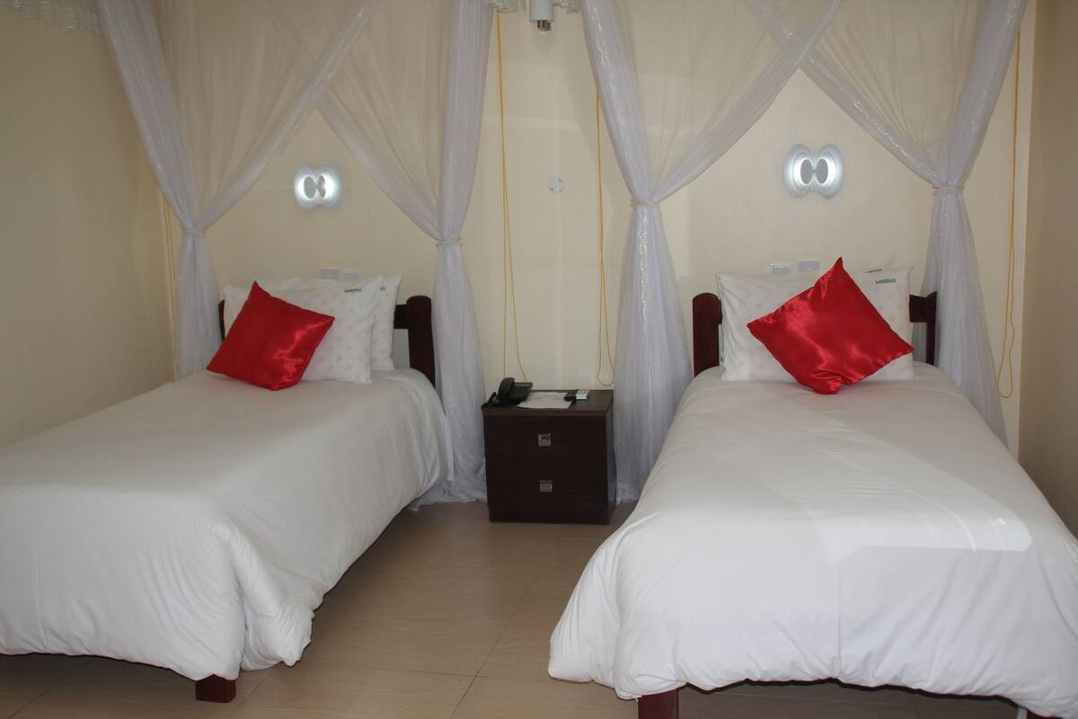 Two beds with white bedding and red pillows, a nightstand between them, and a canopy over the beds.