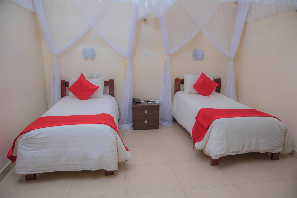 A hotel room with two beds, each with a red pillow and a white bedspread. There is a nightstand between the beds with a phone and a small drawer.