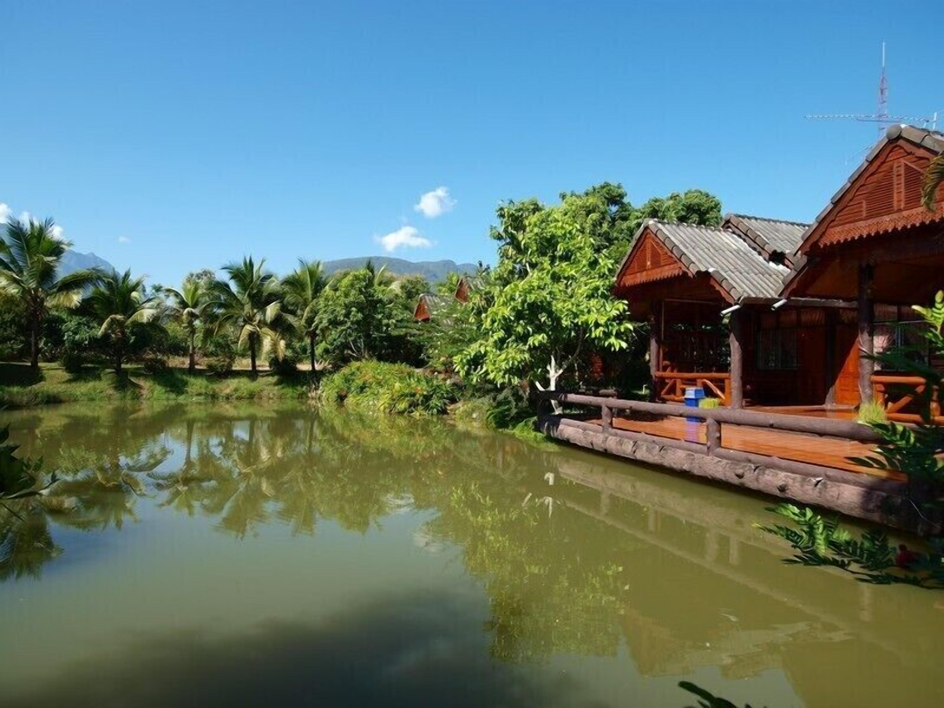 A traditional wooden house with a thatched roof, situated beside a calm pond surrounded by lush greenery and palm trees.