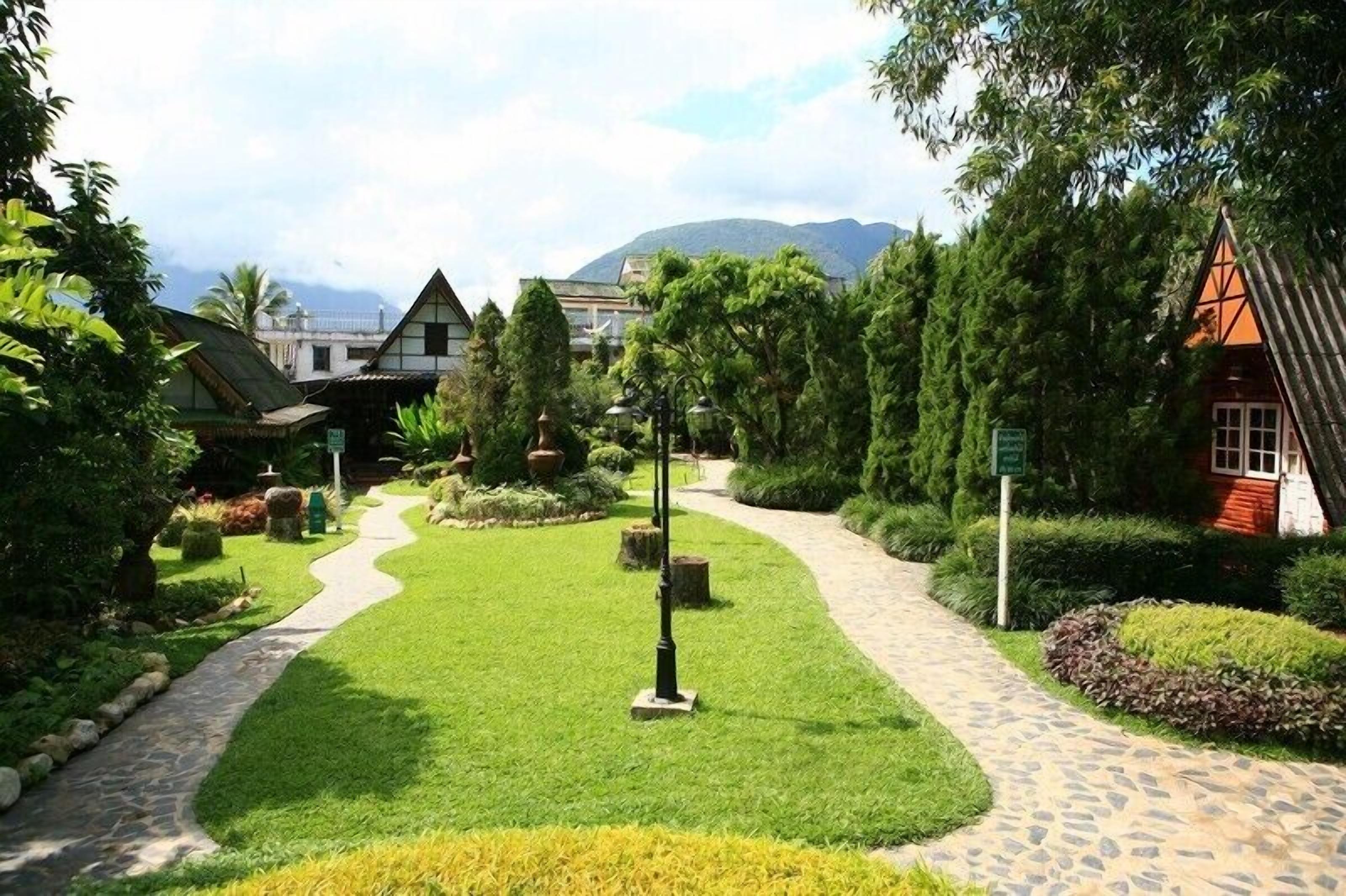 Un sendero en un jardín conduce a una casa tradicional con techo rojo, rodeada de vegetación exuberante y una montaña al fondo.