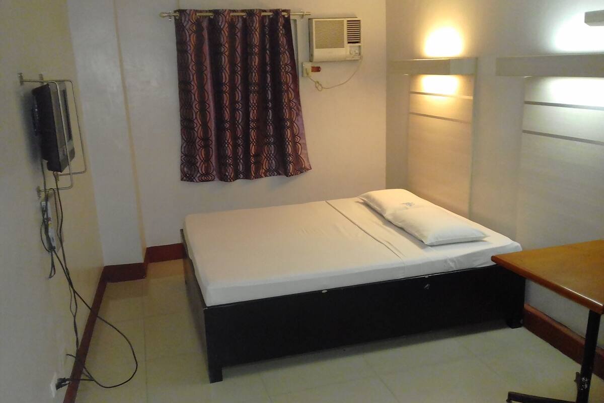 A small, neatly made bed with a white mattress and a single pillow, a wall-mounted air conditioner, and a plain wall with a light switch.