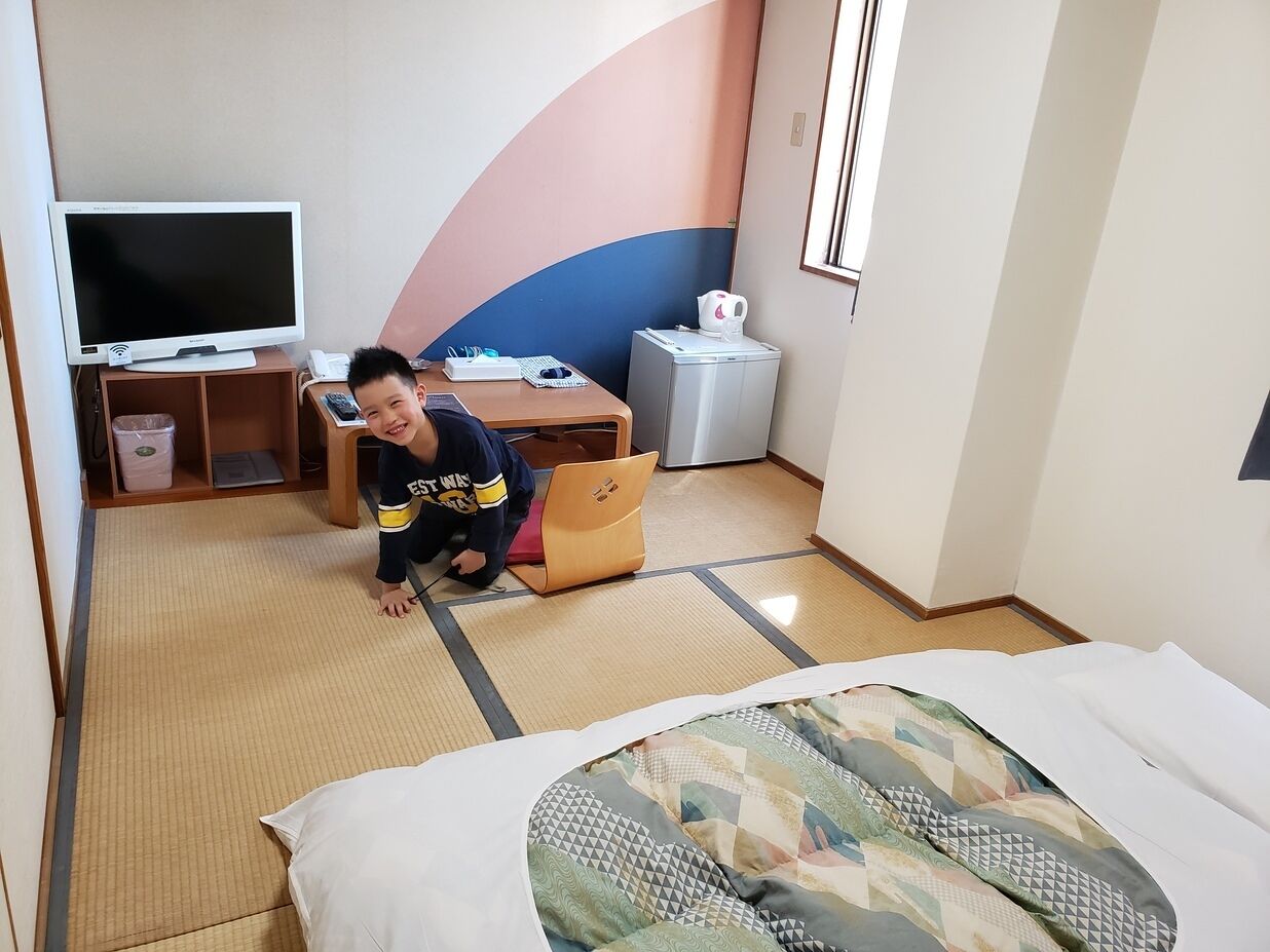 A hotel room with tatami flooring, a bed with a patterned bedspread, a desk with a laptop, a television, and a small refrigerator.