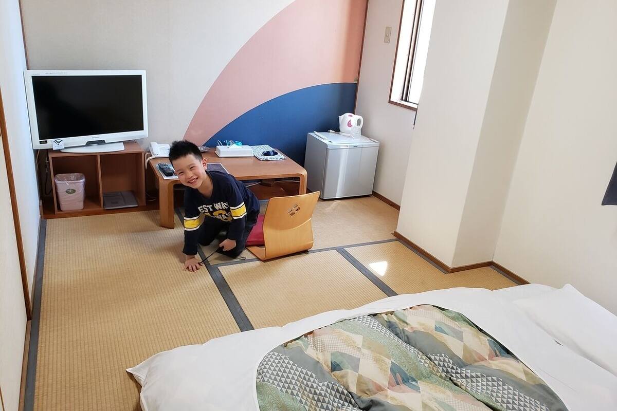 A hotel room with tatami flooring, a bed with a patterned bedspread, a desk with a laptop, a television, and a small refrigerator.