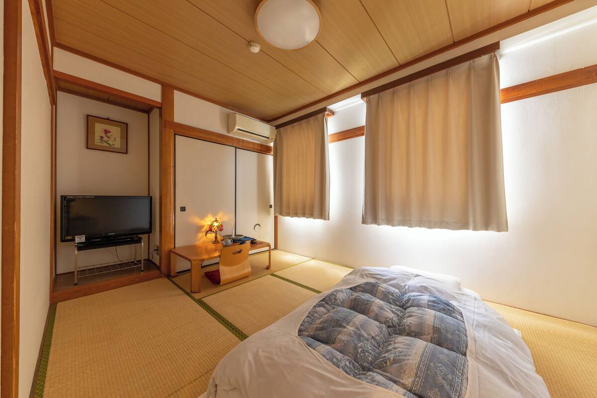 A traditional Japanese-style room with tatami flooring, a bed with a patterned blanket, a television, a desk with a lamp, and a sliding door.