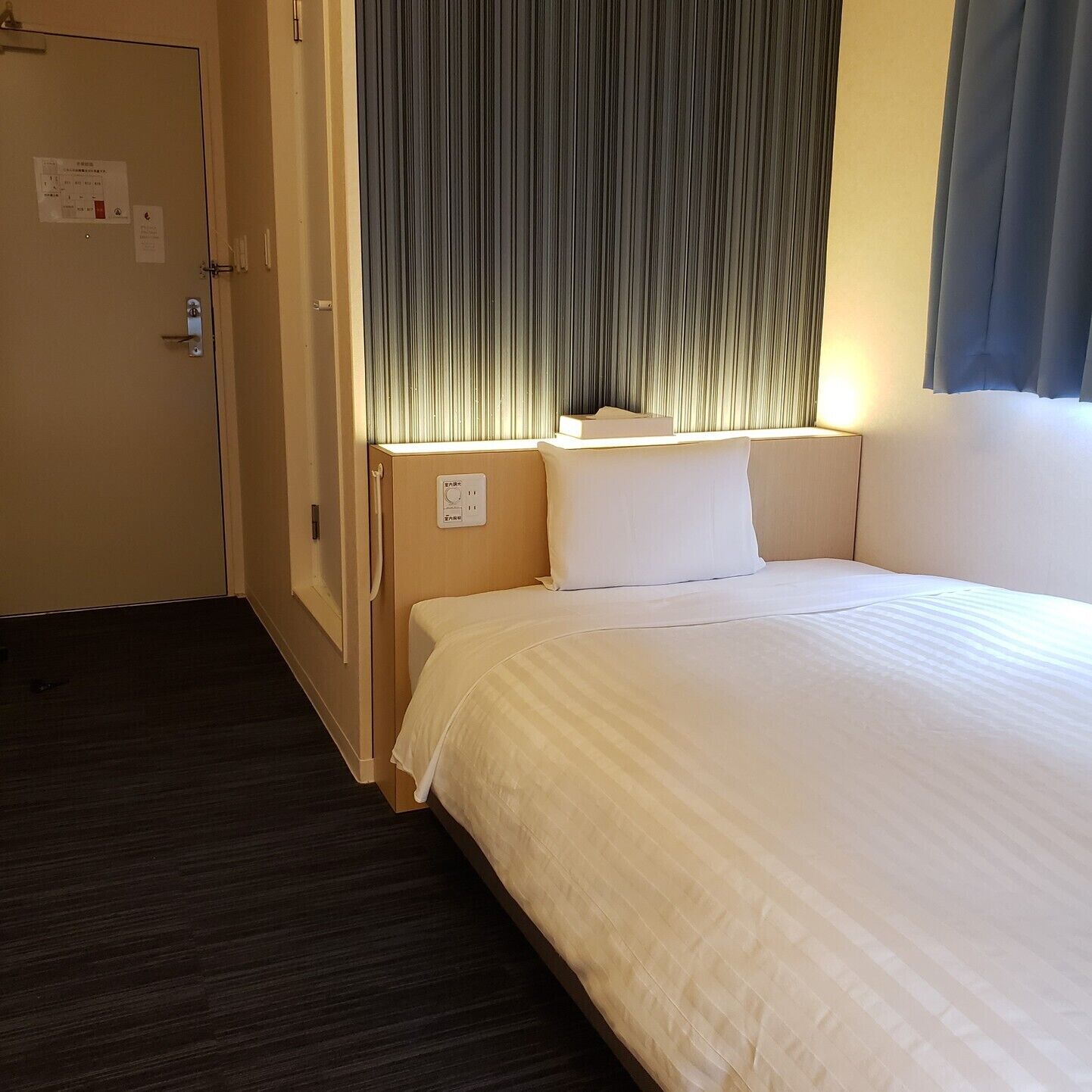 A hotel room with a bed, a nightstand, a window with curtains, a door, and a wall-mounted light.