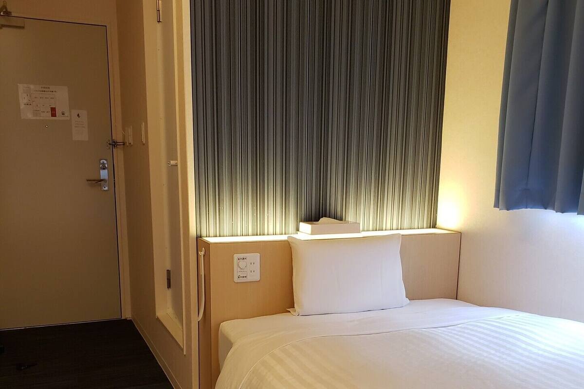 A hotel room with a bed, a nightstand, a window with curtains, a door, and a wall-mounted light.