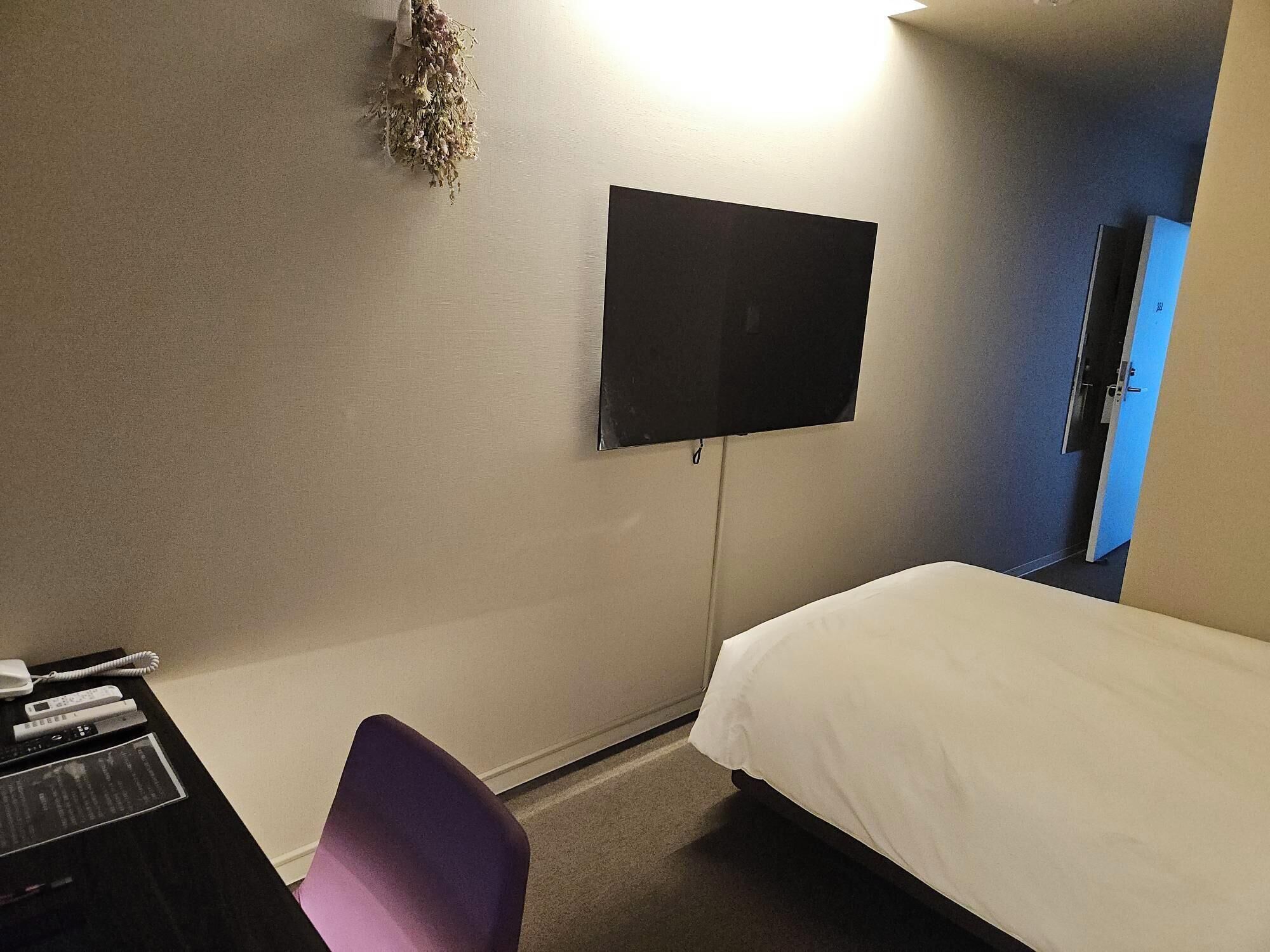 Habitació d'hotel amb un llit, una taula de treball amb un telèfon, una cadira, un televisor de pantalla plana fixat a la paret i una porta amb marc blau.