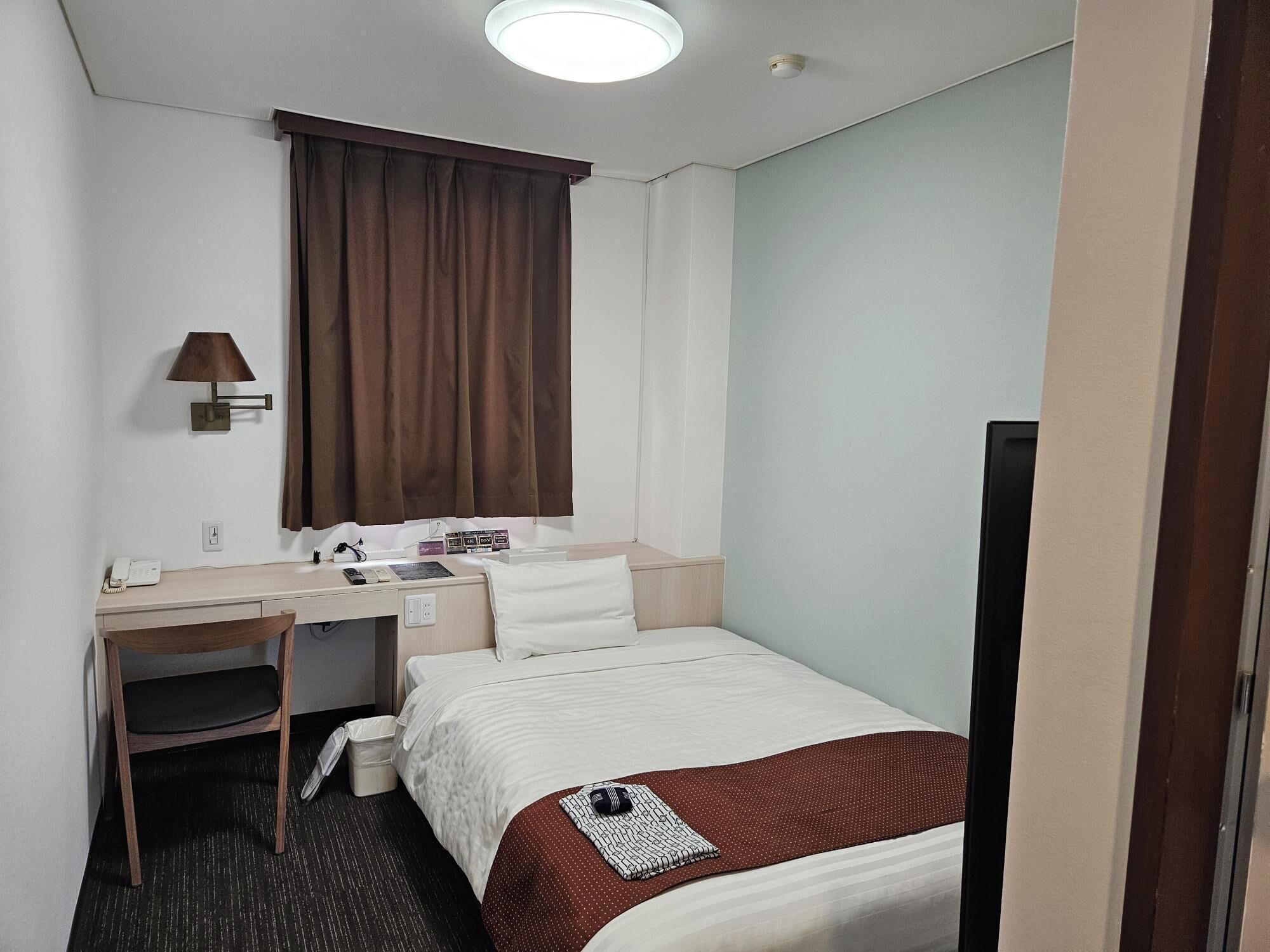 A hotel room with a bed, a desk, a chair, a lamp, a window with curtains, and a television.