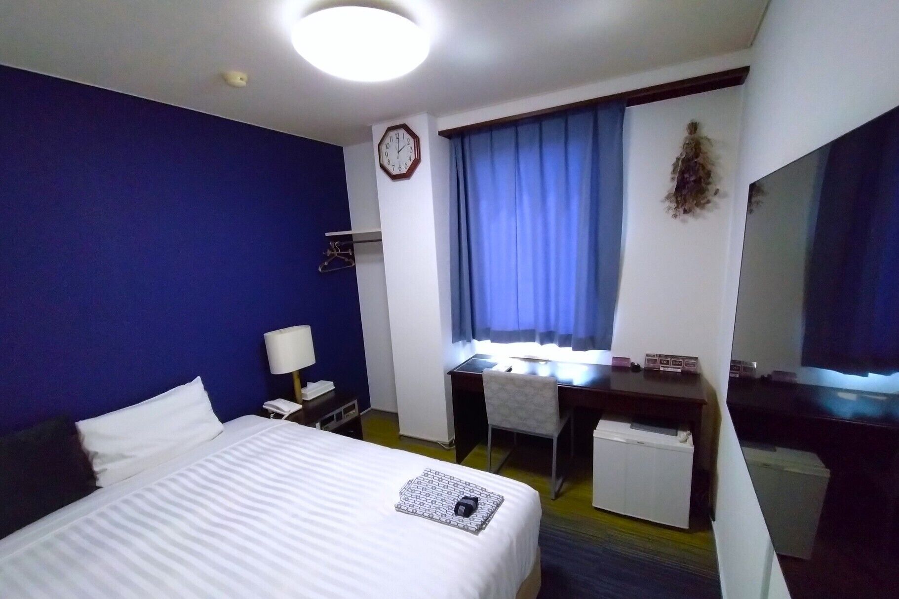A hotel room with a bed, desk, chair, and a clock on the wall.