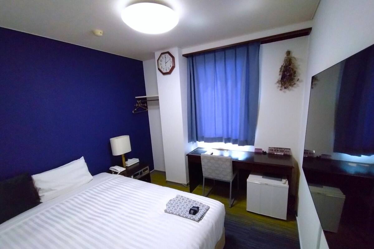 A hotel room with a bed, desk, chair, and a clock on the wall.