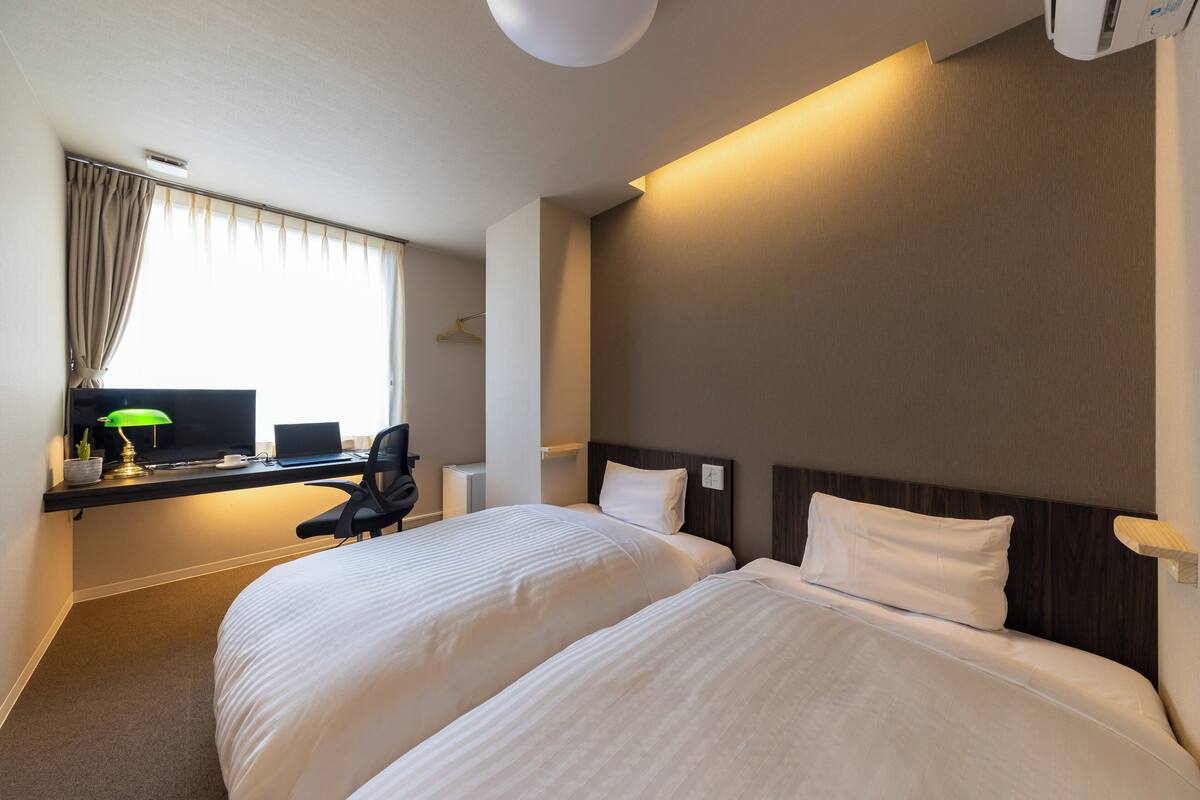 A hotel room with two beds, a desk with a computer, a lamp, and a window with curtains.
