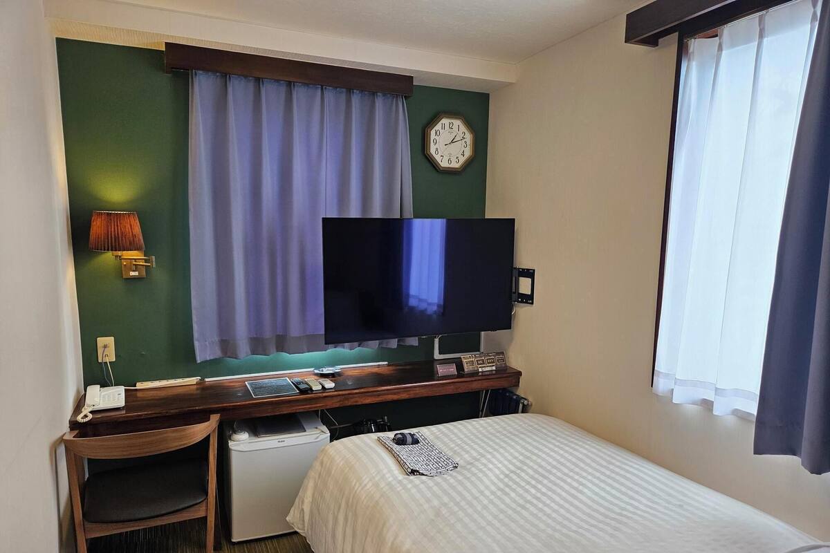 A hotel room with a bed, a desk with a computer, a television, a clock, and a window with curtains.