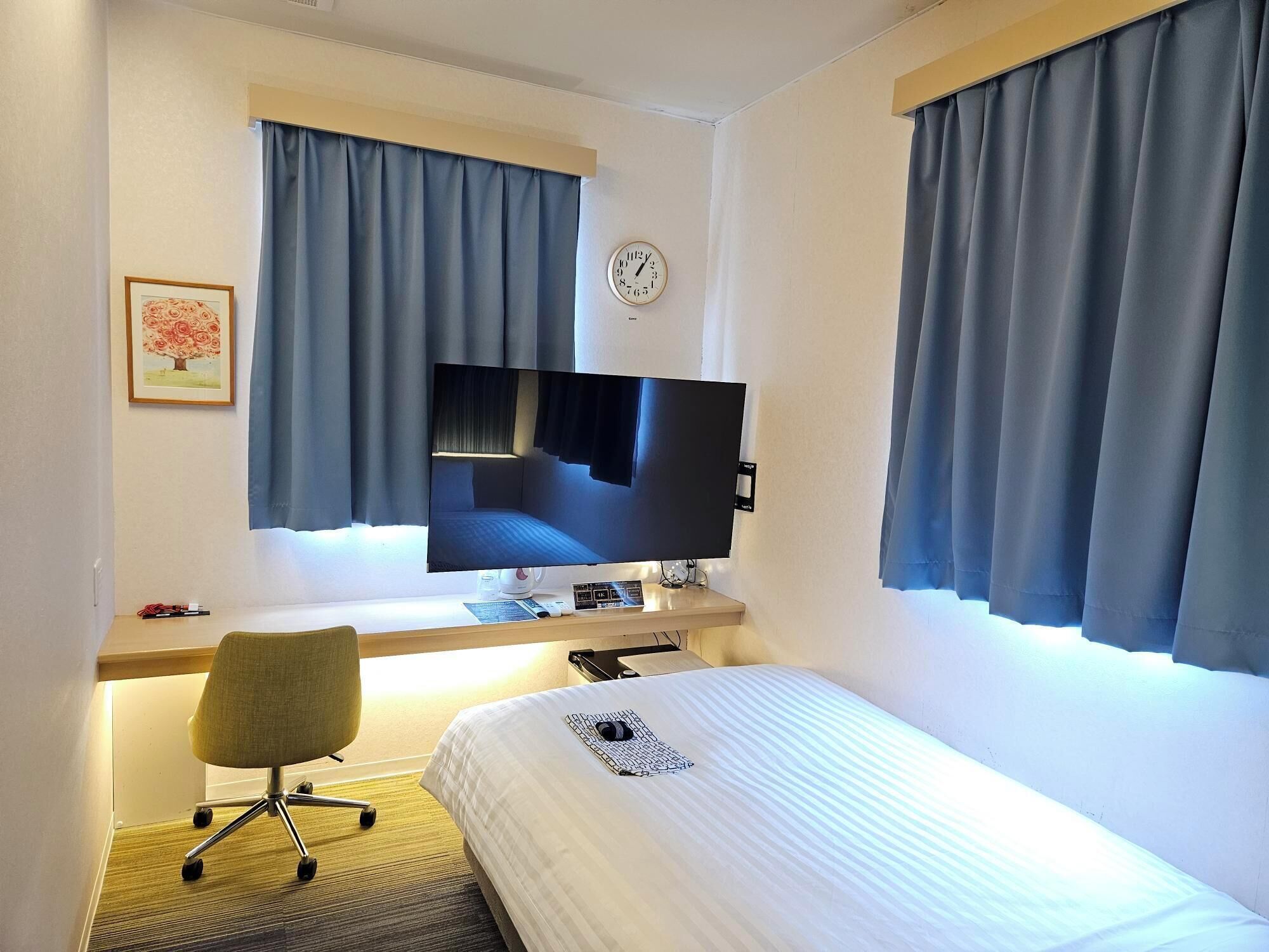 A hotel room with a bed, a desk with a computer, a TV, and a clock on the wall.