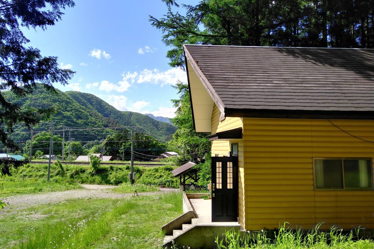 Un bâtiment jaune, surmonté d’un toit sombre, entouré de verdure et de montagnes.