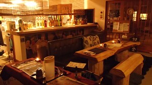 Uma área de bar com iluminação suave, móveis de madeira, um balcão com diversas garrafas e um quadro de cardápio.