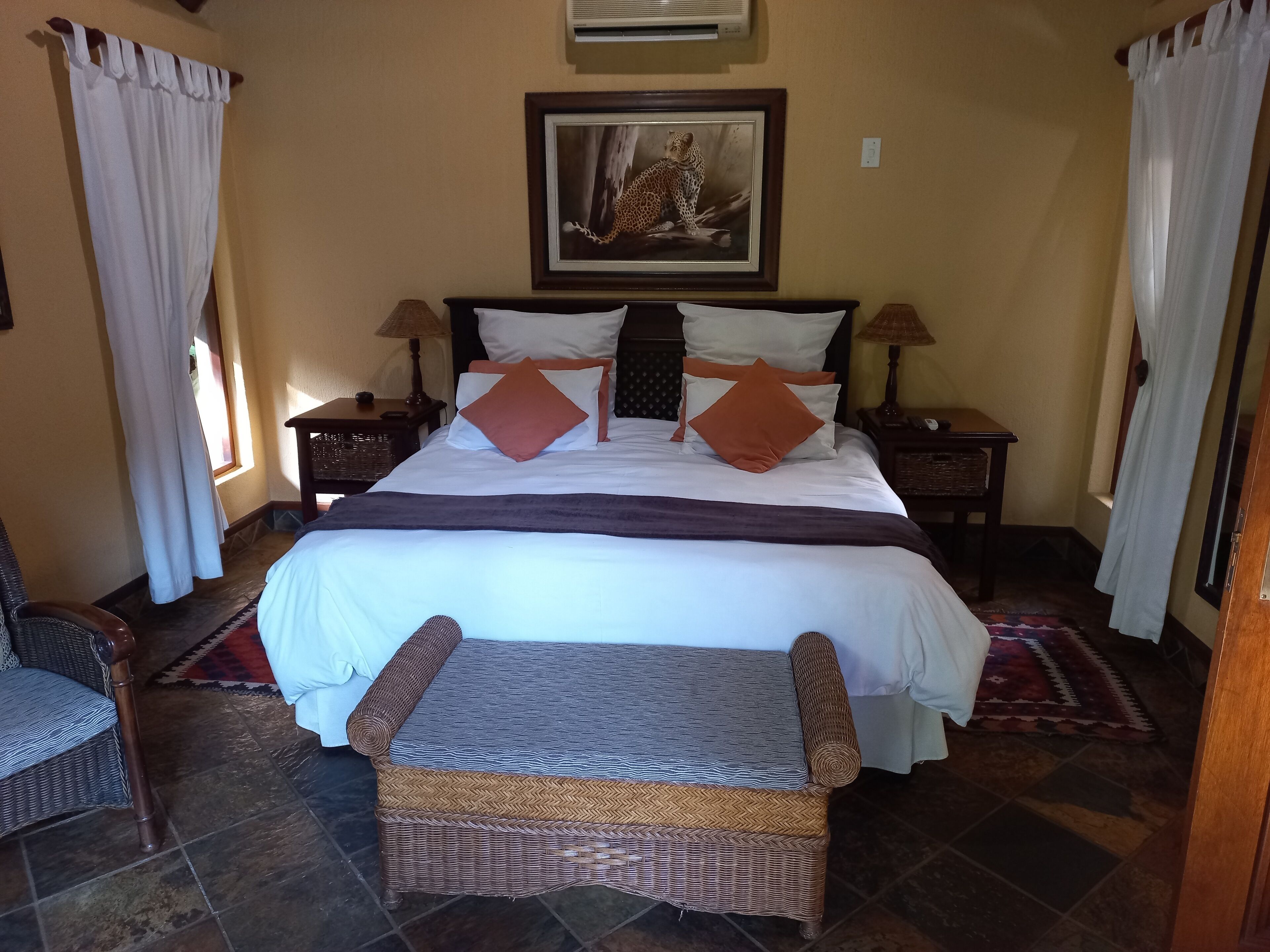 A neatly arranged bedroom with a large bed, two bedside tables, a wicker bench, and a framed picture on the wall.