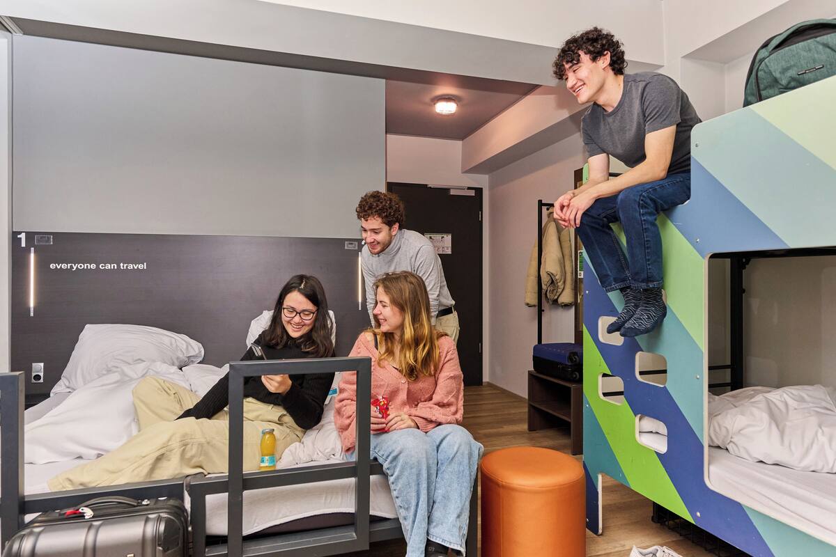 Een slaapzaal met stapelbedden, een bed met een koffer, en een poster aan de muur.