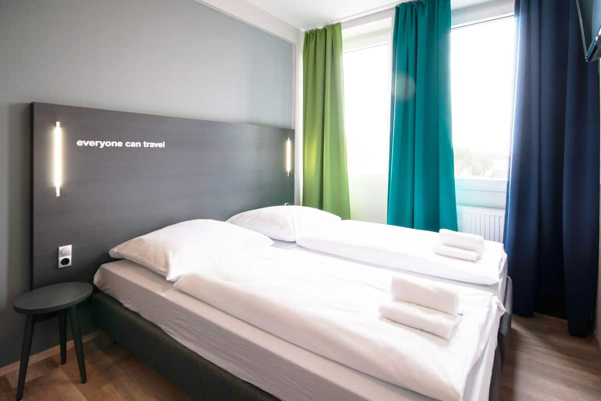 Hotelkamer met twee bedden, een klein krukje, een wandlamp en een raam met gordijnen.