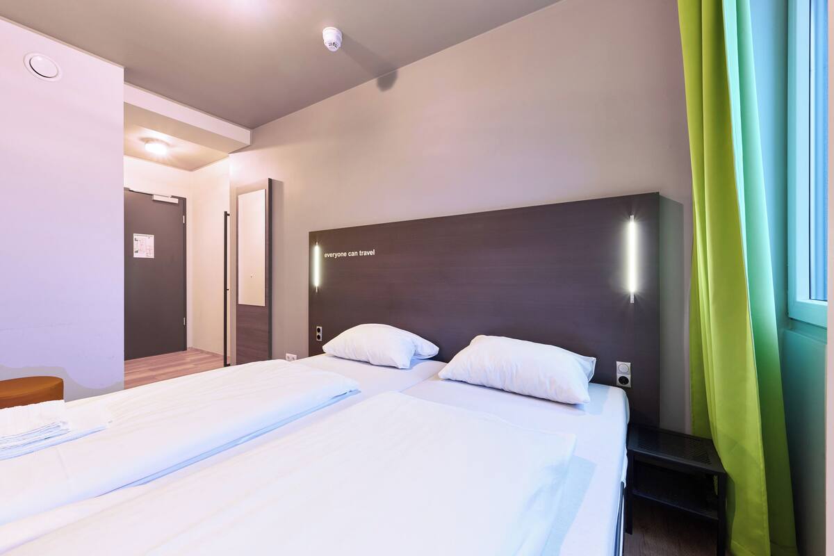 Hotelkamer met twee bedden, een houten hoofdbord, een tafeltje en een raam met groene gordijnen.
