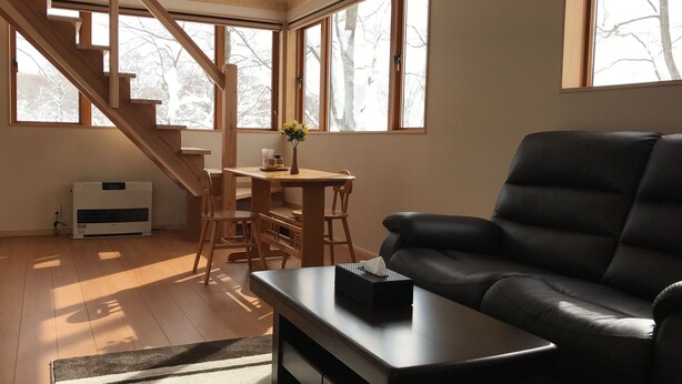 A spacious living room with a black leather sofa, wooden dining table, and chairs. A staircase with wooden railings leads to an upper level. Large windows provide natural light and views of trees. - Gassan Pole Pole Farm (Nishikawa)