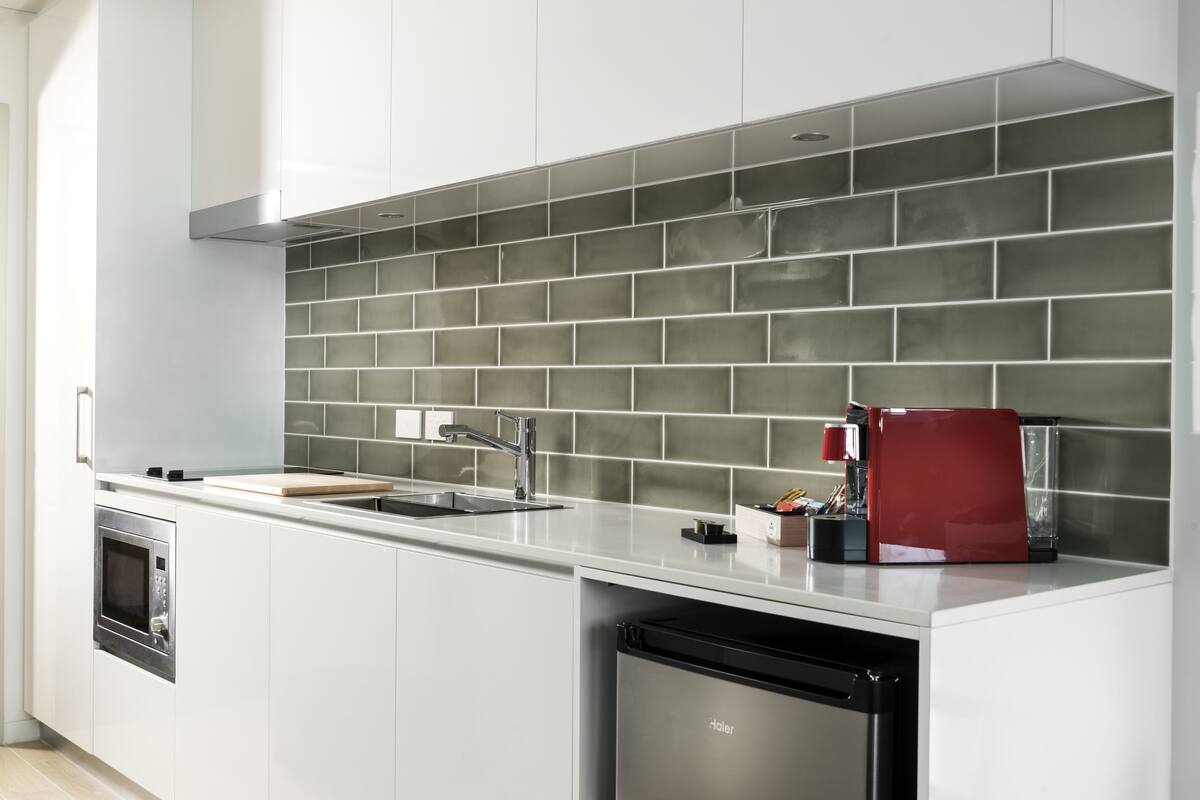 A modern kitchen with white cabinets, a stainless steel oven, a dishwasher, a red coffee machine, and a grey tiled backsplash.