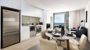 A modern kitchen with a built-in oven, microwave, and refrigerator. There is a dining area with a table and chairs, and a view of the ocean through large windows. - Ramada VetroBlu Scarborough Beach (Scarborough)