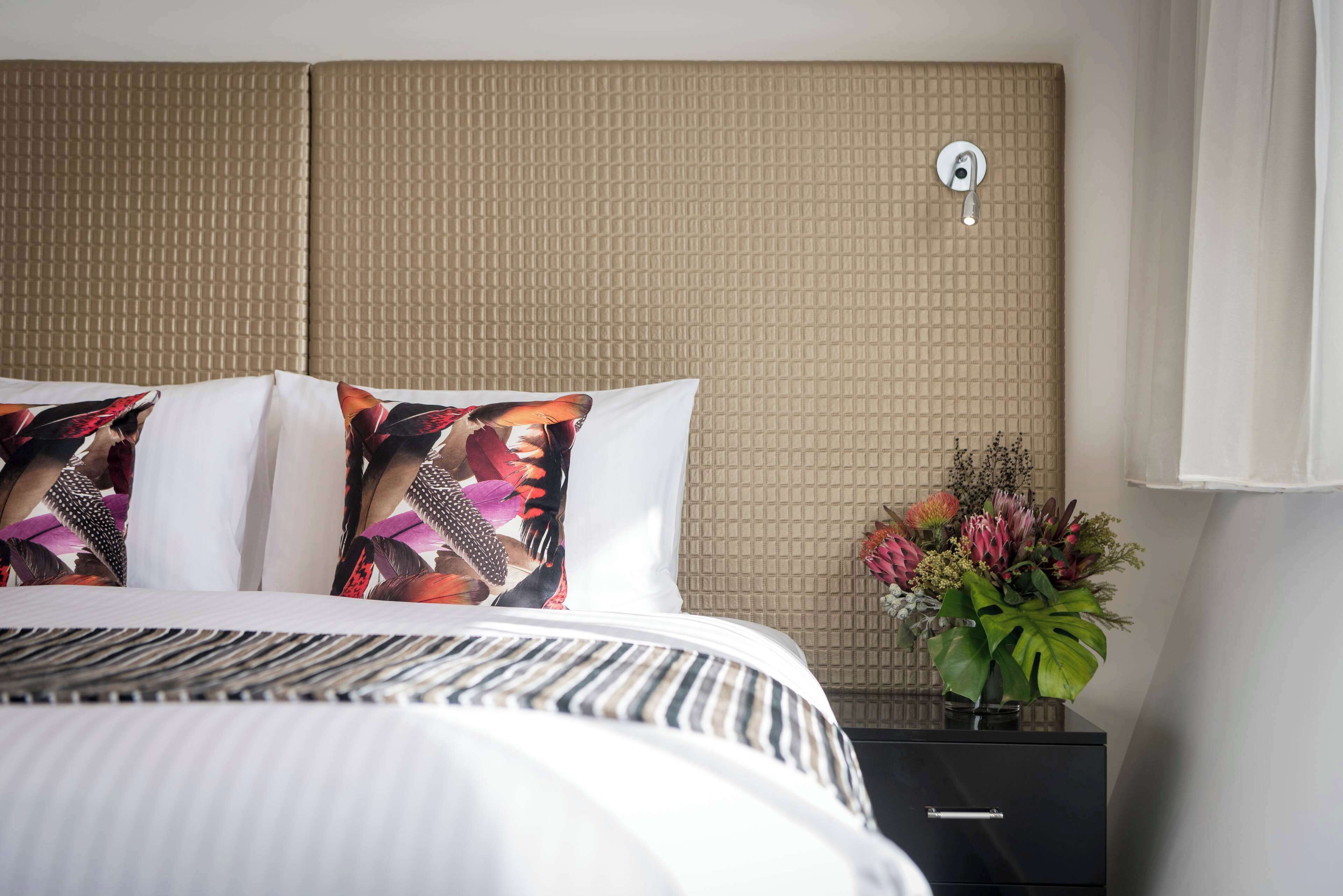 Une chambre d’hôtel avec un grand lit, une tête de lit à motifs, une table de chevet ornée d’un vase de fleurs et un luminaire fixé au mur.