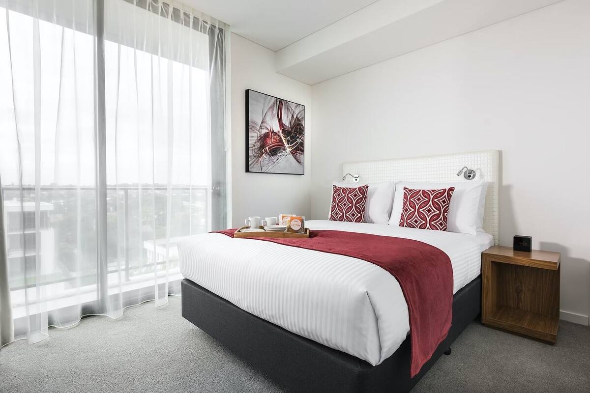 A modern hotel room with a large bed, a bedside table, a window with sheer curtains, and a city view.