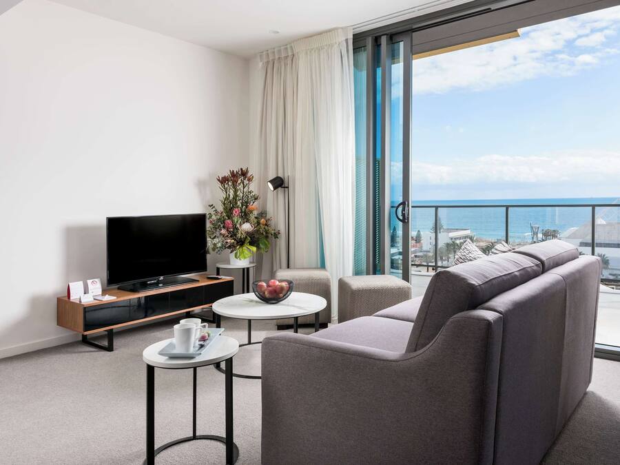 Una habitación de hotel moderna con una televisión de pantalla plana, un sofá, una mesita con un frutero y un balcón con vistas al mar.