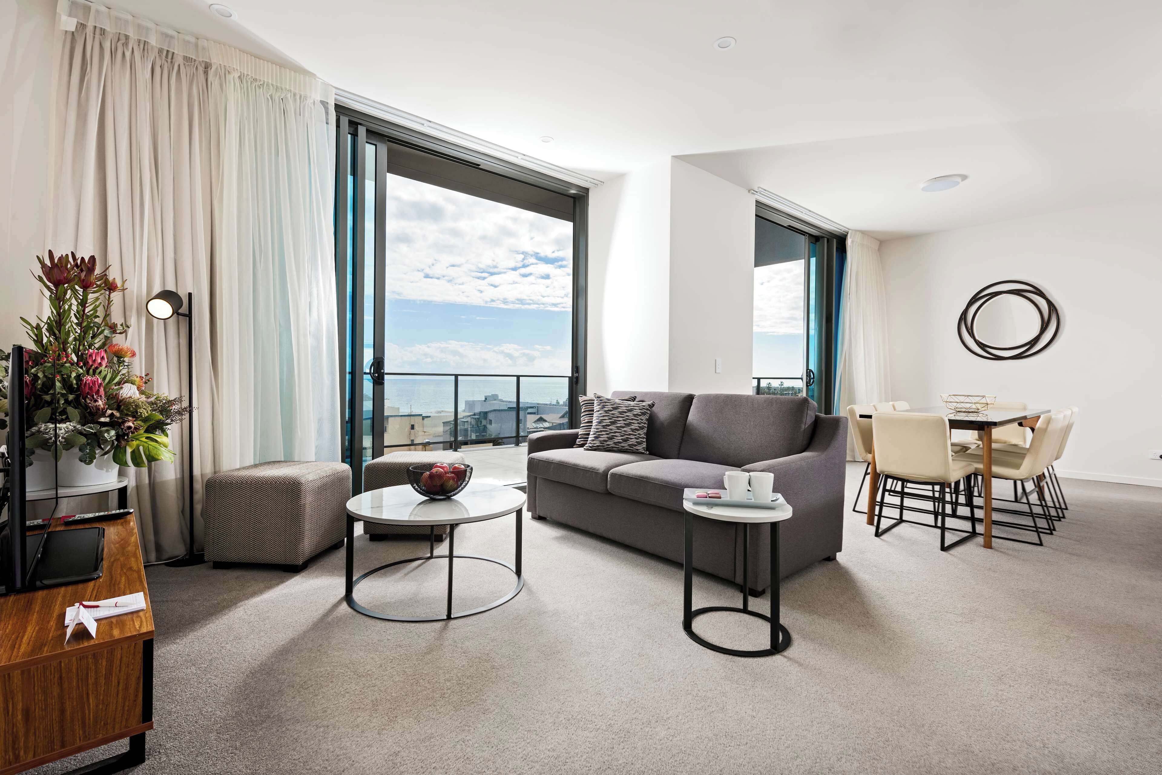 A modern living room with a sofa, coffee table, and dining table. There is a large window with a view of the city.